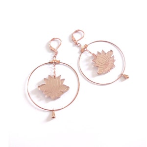 Puede incluir: Pendientes de aro de oro rosa con un colgante de flor de loto. Los pendientes tienen una cadena delicada y una pequeña gota en la parte inferior.