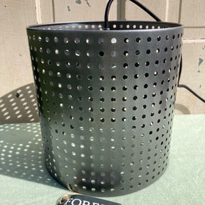 May include: A cylindrical, gray metal pendant light fixture with numerous evenly spaced circular perforations. The light has a black cord and a tag that reads "FORESIDE home garden". The fixture is designed for indoor or outdoor use.