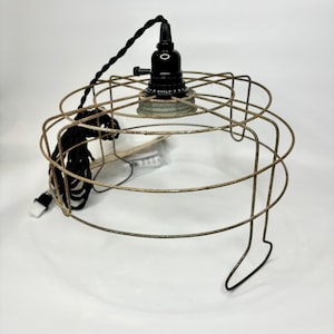 May include: An industrial-style pendant light fixture. It features a black bulb socket, a rusty metal cage shade, and a black braided electrical cord. The cage is circular with a series of horizontal and vertical metal bars.