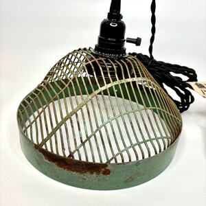 May include: A vintage-style pendant light fixture with a green metal cage shade. The shade has a distressed, aged appearance with rust spots. The fixture includes a black socket and a twisted black electrical cord. The overall design is industrial and rustic.