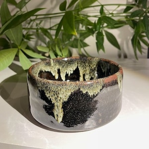 May include: A black ceramic bowl with a brown rim and a drip glaze in shades of green and brown.