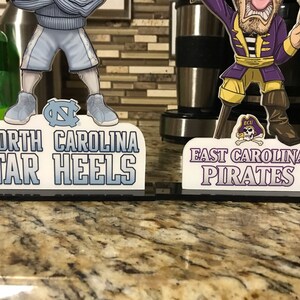 May include: Two cartoon figures stand on black bases with white text. The figure on the left is wearing a light blue uniform with a blue and white logo. The text reads "North Carolina Tar Heels". The figure on the right is wearing a purple and yellow pirate costume with a skull and crossbones logo. The text reads "East Carolina Pirates".