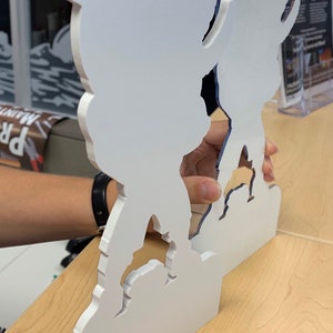 May include: Two white, cutout, cartoon figures stand on a wooden surface. The figures are facing each other and are holding hands. The figure on the left is a person with a backpack. The figure on the right is a person with a hat. The figures are cut out of a white material and have a blue outline.