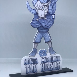 May include: A blue and white 3D cutout of the North Carolina Tar Heels mascot, a ram with a blue sweater and shorts, standing on a black base. The base has the words "North Carolina Tar Heels" printed on it.