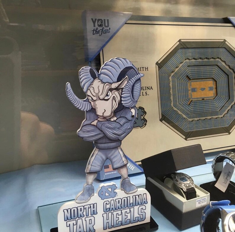 May include: A blue and white figurine of a ram mascot with the text "North Carolina Tar Heels" on the base. The figurine is standing on a black base with a glass top. The figurine is wearing a blue and white jersey with the number "1" on the front. The figurine is holding a basketball in its right hand. The figurine is standing in front of a framed picture of a basketball court.