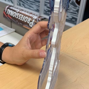 May include: A white and blue 3D printed sign with a whimsical design. The sign features a stylized image of a person with a blue and white hat and a white beard. The sign is made of a thick, durable material and has a smooth, glossy finish.