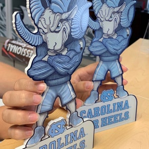 May include: Two blue and white Carolina Tar Heels mascot figurines with the team logo and name on the base. The mascots are standing with their arms crossed and are wearing blue and white uniforms.