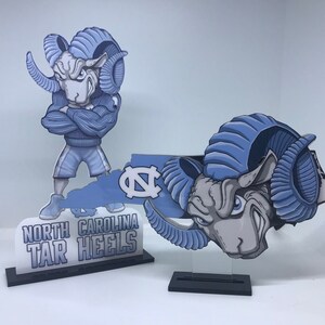 May include: Two blue and gray ram mascot figurines, one standing on a white base with the text "North Carolina Tar Heels", the other on a black base. Both figurines are in the shape of a ram's head with blue horns and a gray face.