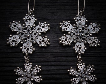 Può includere: Un paio di orecchini a forma di fiocco di neve in argento con un design delicato e intricato. Ogni orecchino presenta due fiocchi di neve, uno più grande dell'altro, collegati da una piccola catena.