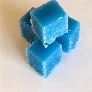 May include: Four blue sugar scrub cubes stacked on top of each other. The cubes are made with a coarse sugar and have a light blue color.