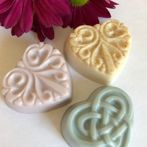 May include: Three heart-shaped soaps with different designs. The soap on the left is lavender with a floral design. The soap in the center is beige with a swirl design. The soap on the right is light blue with a Celtic knot design.