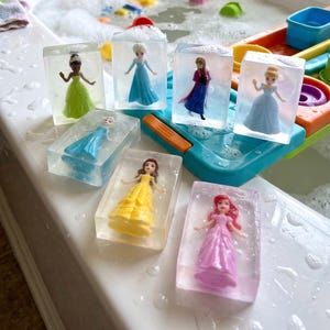 May include: Six rectangular clear soap bars, each containing a small plastic princess figurine. The figures include characters in green, blue, yellow, and pink dresses, embedded within the transparent soap blocks, resting on a white surface.