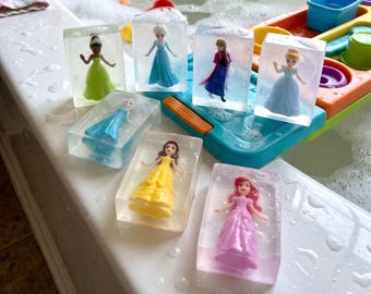 May include: Six rectangular clear soap bars, each containing a small plastic princess figurine. The figures include characters in green, blue, yellow, and pink dresses, embedded within the transparent soap blocks, resting on a white surface.