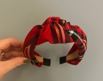 May include: A red plaid fabric headband with a knotted top. The headband features a classic tartan pattern in red, black, yellow, and white. The ends are finished with black fabric.