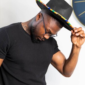 K&ouml;nnte beinhalten: Ein schwarzer Fedora-Hut mit einem bunten Band. Der Hut wird von einer Person gehalten, die ein schwarzes T-Shirt und eine Brille tr&auml;gt.