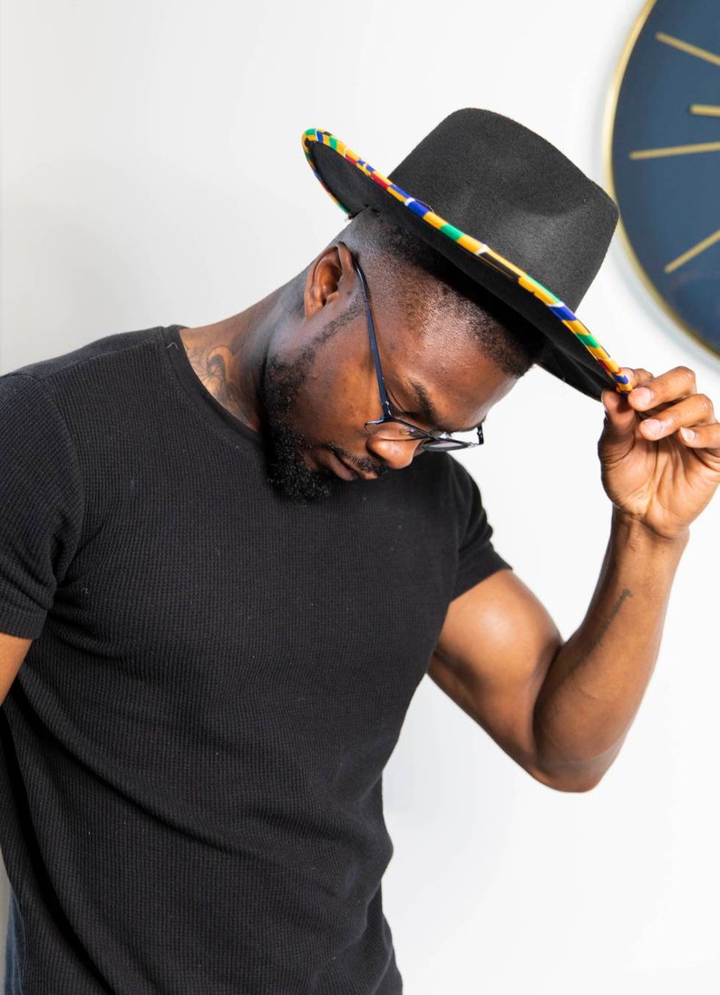 K&ouml;nnte beinhalten: Ein schwarzer Fedora-Hut mit einem bunten Band. Der Hut wird von einer Person gehalten, die ein schwarzes T-Shirt und eine Brille tr&auml;gt.
