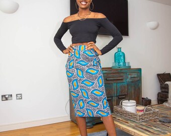 May include: A woman wearing a black off-the-shoulder top, a blue and white patterned skirt, and clear heeled sandals. She is standing on a gray rug in front of a wooden table.