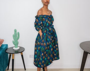 May include: A woman wearing a blue off-the-shoulder dress with a green and gold leaf pattern. The dress has a cinched waist and a flowing skirt. She is wearing black heels.