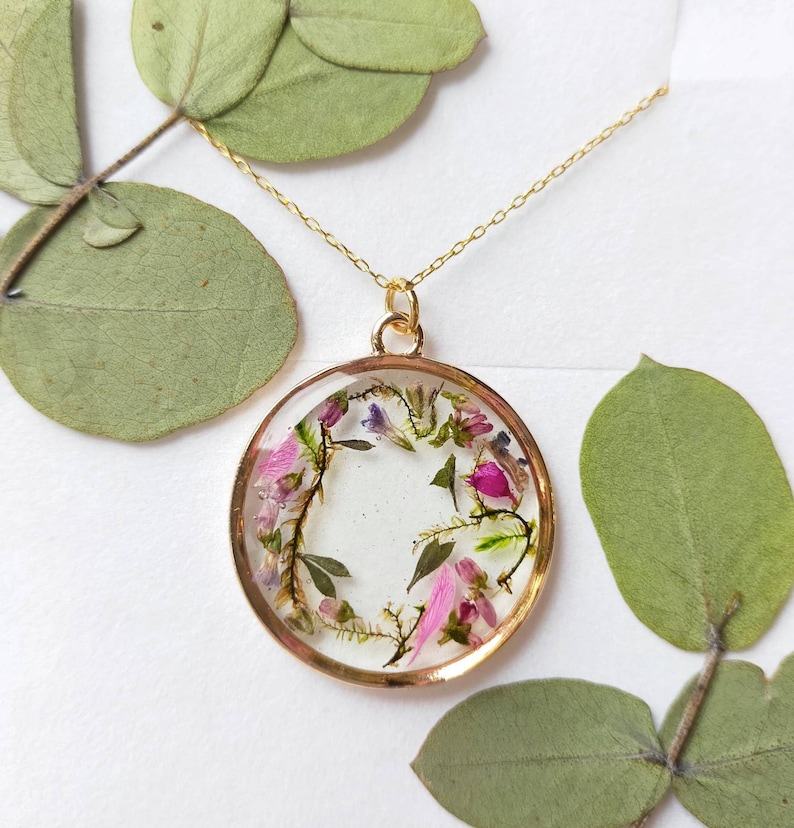 May include: A gold chain necklace with a round pendant featuring a delicate arrangement of pressed pink and purple flowers and green leaves encased in clear resin.