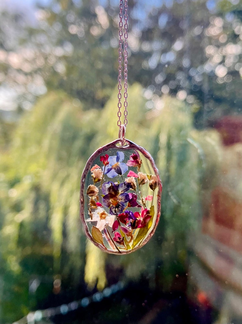 Pode incluir: Um colar de corrente de prata com um pingente oval transparente contendo flores prensadas. As flores s&atilde;o roxas, azuis, cor-de-rosa e brancas.