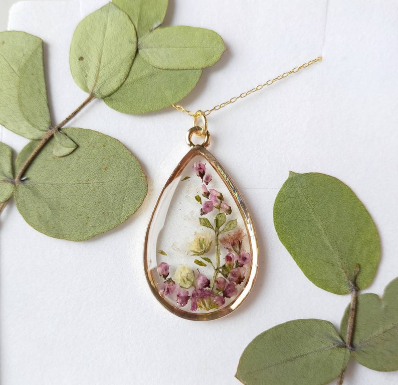 May include: A gold-toned pendant necklace with a teardrop-shaped resin charm. The charm encases small, dried pink and white flowers. The necklace is displayed on a white surface with green leaves.