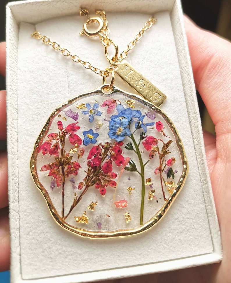 以下が含まれることがあります： 金色のネックレスに、押し花と金箔で飾られた丸いペンダントがついたもの。ペンダントは透明で、ピンク、青、緑の花が見える。ネックレスには、「H & S」と刻まれた小さな金色のタグがついている。