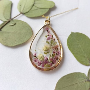 May include: A gold-colored pendant necklace with a teardrop-shaped resin charm. The charm contains dried pink and white flowers. The necklace is displayed on a white surface with green leaves in the background.