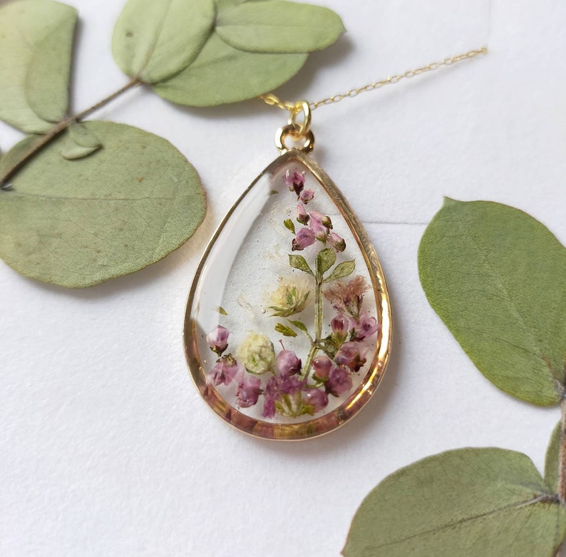 May include: A gold-colored pendant necklace with a teardrop-shaped resin charm. The charm contains dried pink and white flowers. The necklace is displayed on a white surface with green leaves in the background.