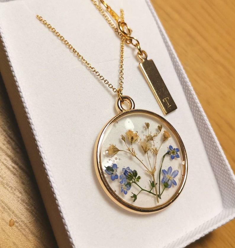 K&ouml;nnte beinhalten: Eine goldene Halskette mit einem runden Anh&auml;nger, der gepresste blaue und wei&szlig;e Blumen in klarem Harz enth&auml;lt. Der Anh&auml;nger ist an einer zarten goldenen Kette befestigt. Ein kleines goldenes Schild mit dem Buchstaben "T" ist ebenfalls an der Kette befestigt.