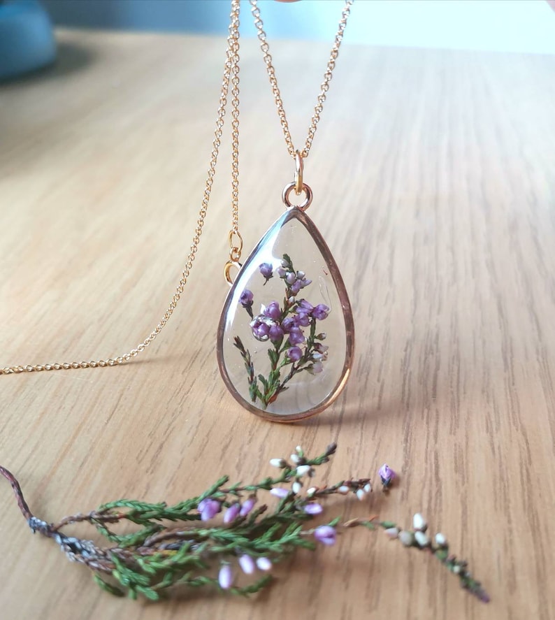 May include: A gold chain necklace with a teardrop-shaped pendant. The pendant is filled with clear resin and contains a small sprig of purple flowers.