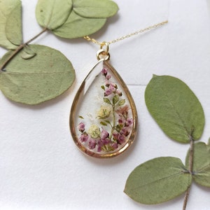 May include: A gold-toned pendant necklace with a teardrop-shaped resin charm. The charm encases small, dried purple and white flowers. The necklace is displayed on a white surface, with green leaves in the background.