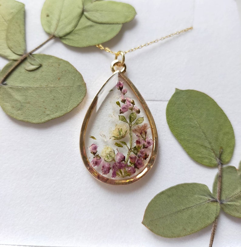May include: A gold-toned pendant necklace with a teardrop-shaped resin charm. The charm encases small, dried purple and white flowers. The necklace is displayed on a white surface, with green leaves in the background.