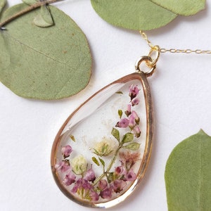 May include: A teardrop-shaped pendant necklace with a gold-colored frame. The clear resin pendant contains dried pink and white flowers and green leaves. The necklace chain is also gold-colored. The background includes green leaves.