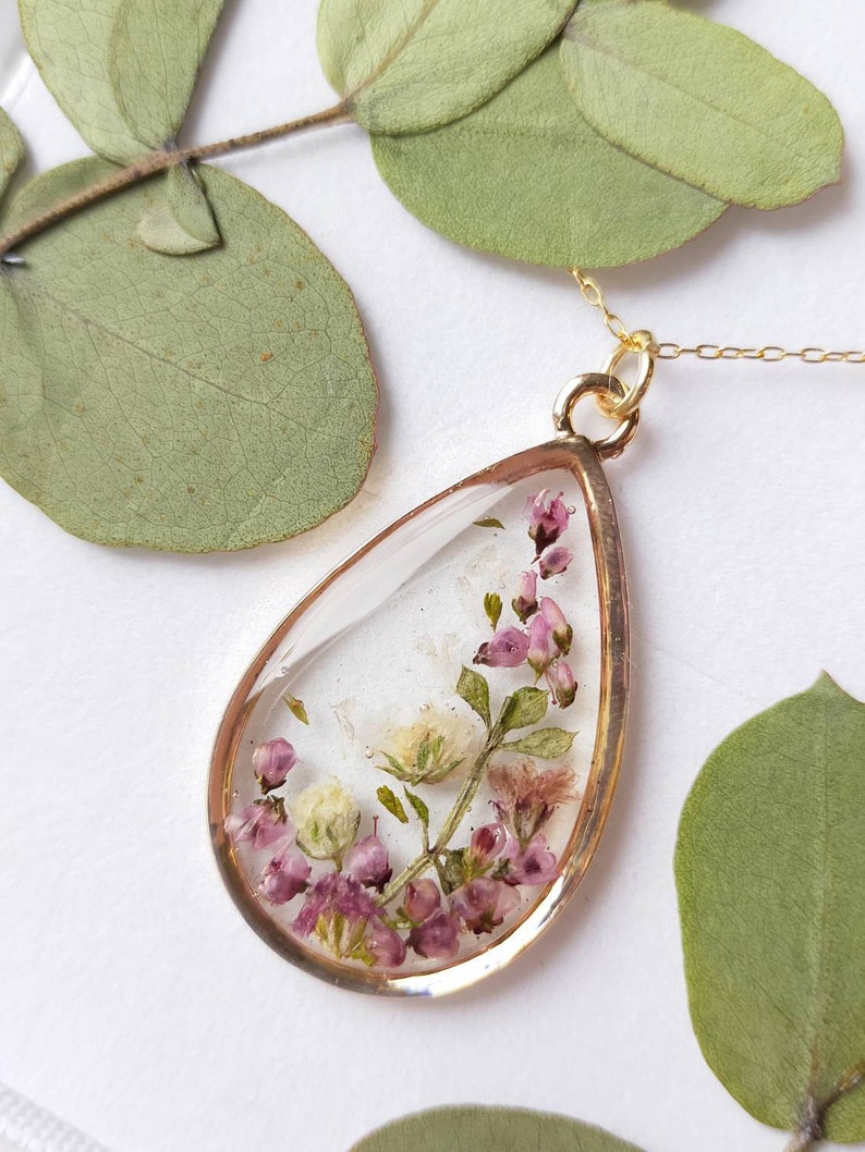 May include: A teardrop-shaped pendant necklace with a gold-colored frame. The clear resin pendant contains dried pink and white flowers and green leaves. The necklace chain is also gold-colored. The background includes green leaves.