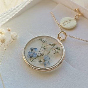 K&ouml;nnte beinhalten: Eine goldene Halskette mit einem runden Anh&auml;nger, der mit einem gepressten Blumenmuster verziert ist. Der Anh&auml;nger enth&auml;lt blaue und wei&szlig;e Blumen. Die Halskette hat auch einen kleinen runden Anh&auml;nger mit dem Wort "Liebe" eingraviert.