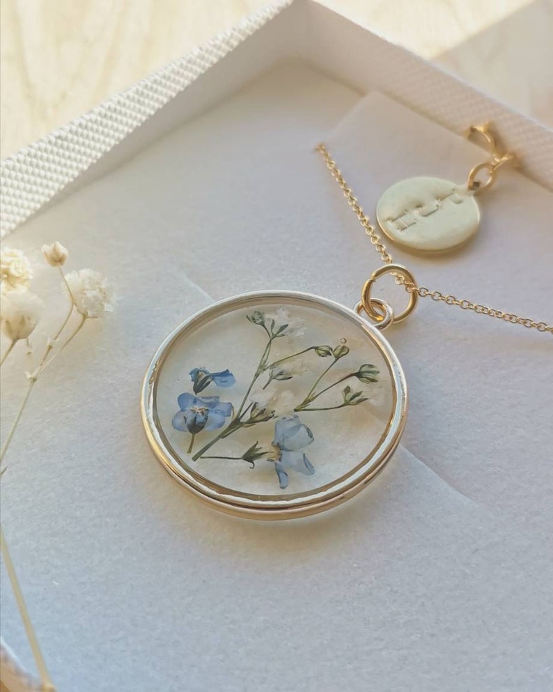 K&ouml;nnte beinhalten: Eine goldene Halskette mit einem runden Anh&auml;nger, der mit einem gepressten Blumenmuster verziert ist. Der Anh&auml;nger enth&auml;lt blaue und wei&szlig;e Blumen. Die Halskette hat auch einen kleinen runden Anh&auml;nger mit dem Wort "Liebe" eingraviert.