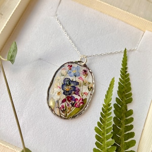 Pode incluir: Um colar de corrente de prata com um pingente oval que apresenta flores prensadas em tons de azul, roxo, rosa e branco. O pingente tem uma borda prateada.