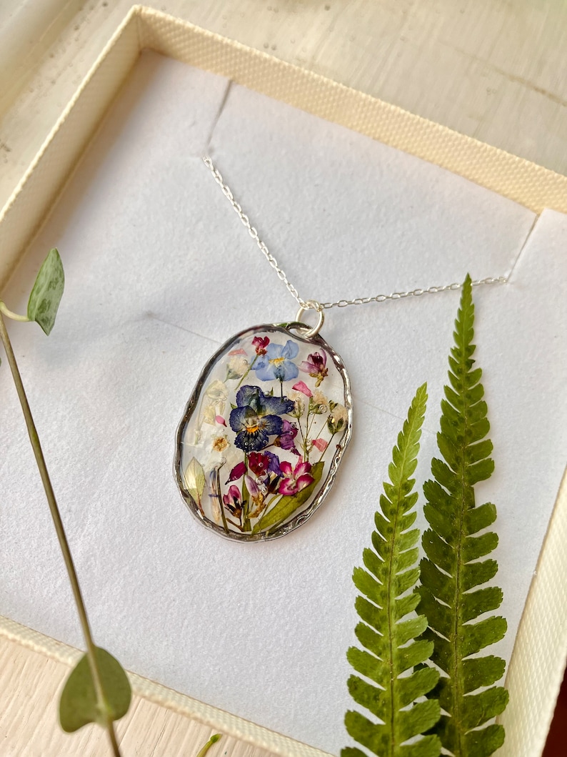 Pode incluir: Um colar de corrente de prata com um pingente oval que apresenta flores prensadas em tons de azul, roxo, rosa e branco. O pingente tem uma borda prateada.