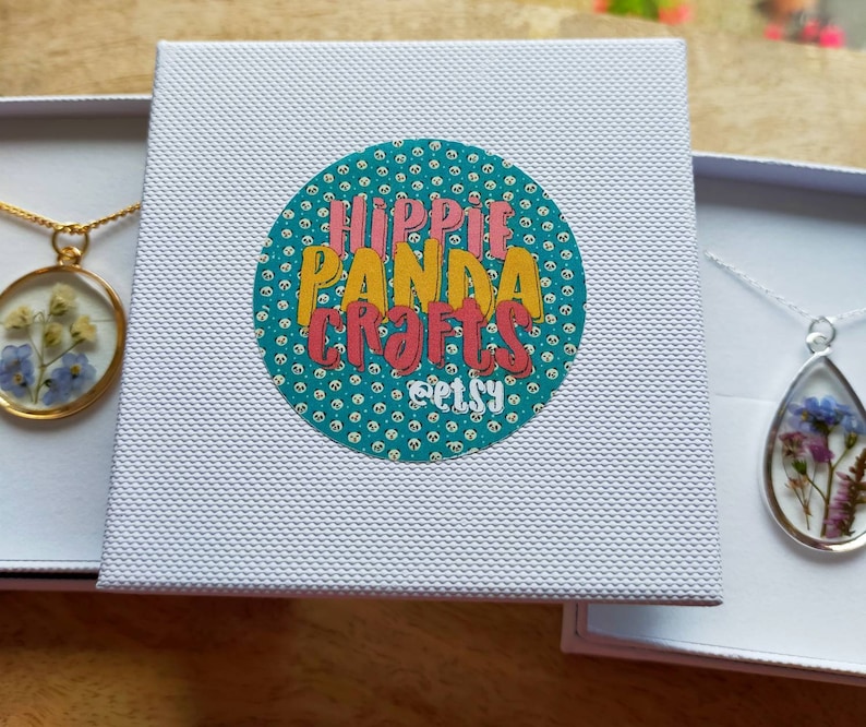 K&ouml;nnte beinhalten: Zwei wei&szlig;e Geschenkboxen mit ge&ouml;ffneten Deckeln, jede mit einer zarten Halskette mit einem Glasanh&auml;nger gef&uuml;llt mit gepressten Blumen. Die Box auf der linken Seite hat eine goldene Kette und einen runden Anh&auml;nger mit blauen Blumen. Die Box auf der rechten Seite hat eine silberne Kette und einen tropfenf&ouml;rmigen Anh&auml;nger mit rosa und blauen Blumen. Die Box auf der linken Seite hat auch einen Aufkleber mit dem Text "Hippie Panda Crafts Etsy" in einem bunten Kreis.