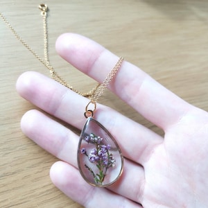 May include: A gold chain necklace with a teardrop-shaped pendant. The pendant is filled with clear resin and contains a pressed purple flower.