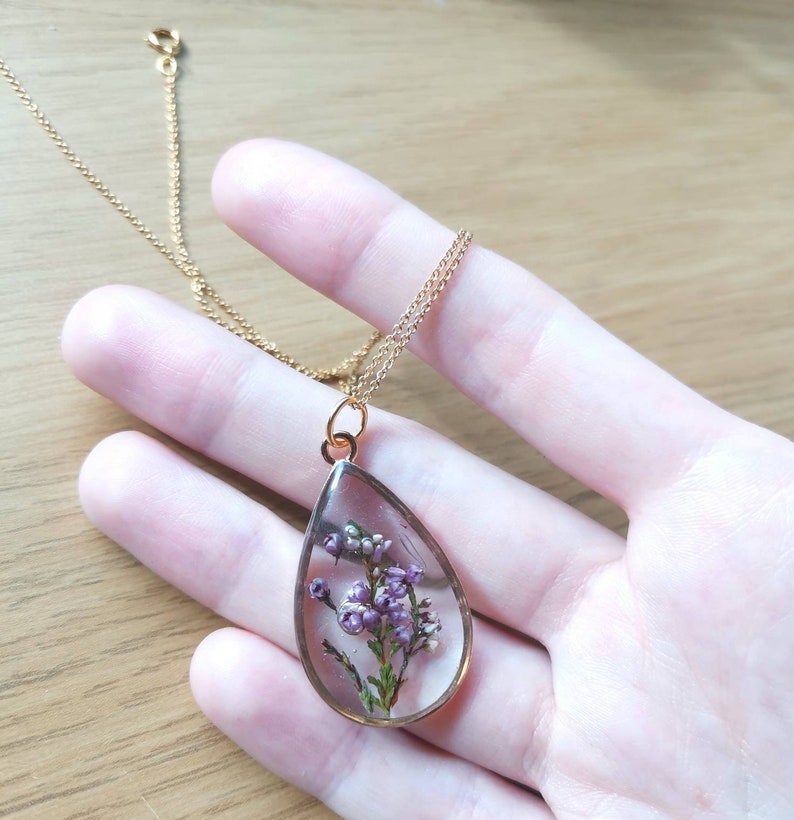 May include: A gold chain necklace with a teardrop-shaped pendant. The pendant is filled with clear resin and contains a pressed purple flower.