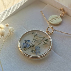 K&ouml;nnte beinhalten: Eine goldene Halskette mit einem runden Anh&auml;nger, der gepresste blaue und wei&szlig;e Blumen in klarem Harz enth&auml;lt. Die Halskette enth&auml;lt auch einen kleinen runden Anh&auml;nger mit dem Wort "Love" eingraviert.