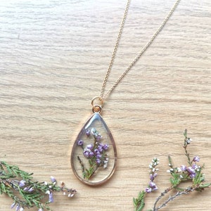 May include: A gold chain necklace with a teardrop-shaped pendant. The pendant is filled with clear resin and contains preserved purple and white flowers.