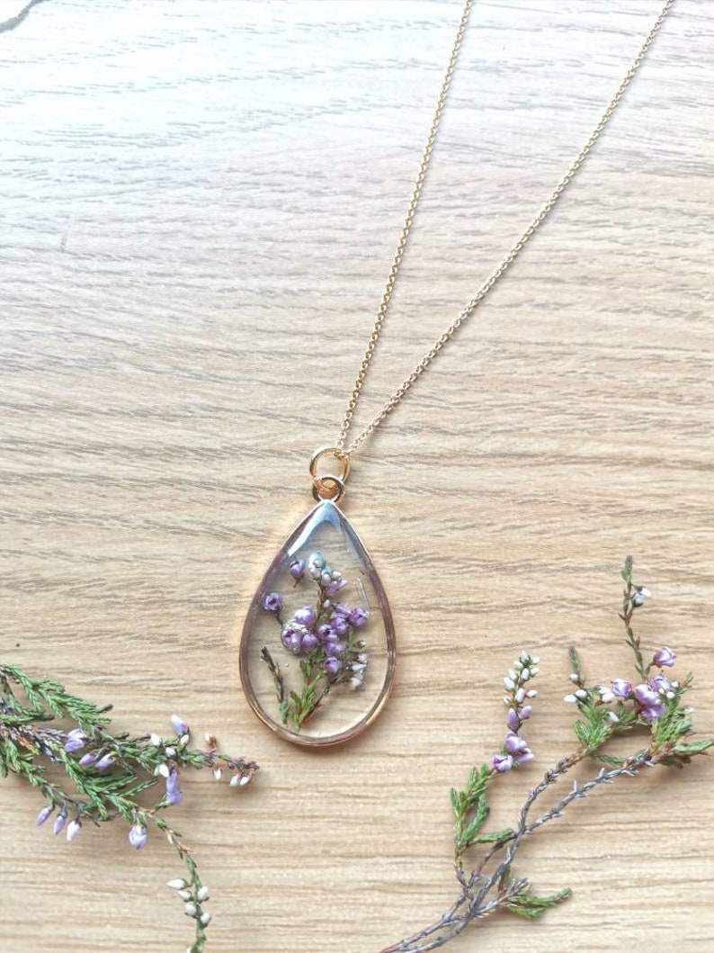 May include: A gold chain necklace with a teardrop-shaped pendant. The pendant is filled with clear resin and contains preserved purple and white flowers.