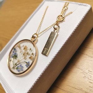K&ouml;nnte beinhalten: Eine goldene Halskette mit einem runden Anh&auml;nger, der gepresste blaue und wei&szlig;e Blumen enth&auml;lt. Der Anh&auml;nger ist an einer zarten goldenen Kette befestigt. Die Halskette enth&auml;lt au&szlig;erdem einen kleinen goldenen Anh&auml;nger mit dem eingravierten Buchstaben "T".