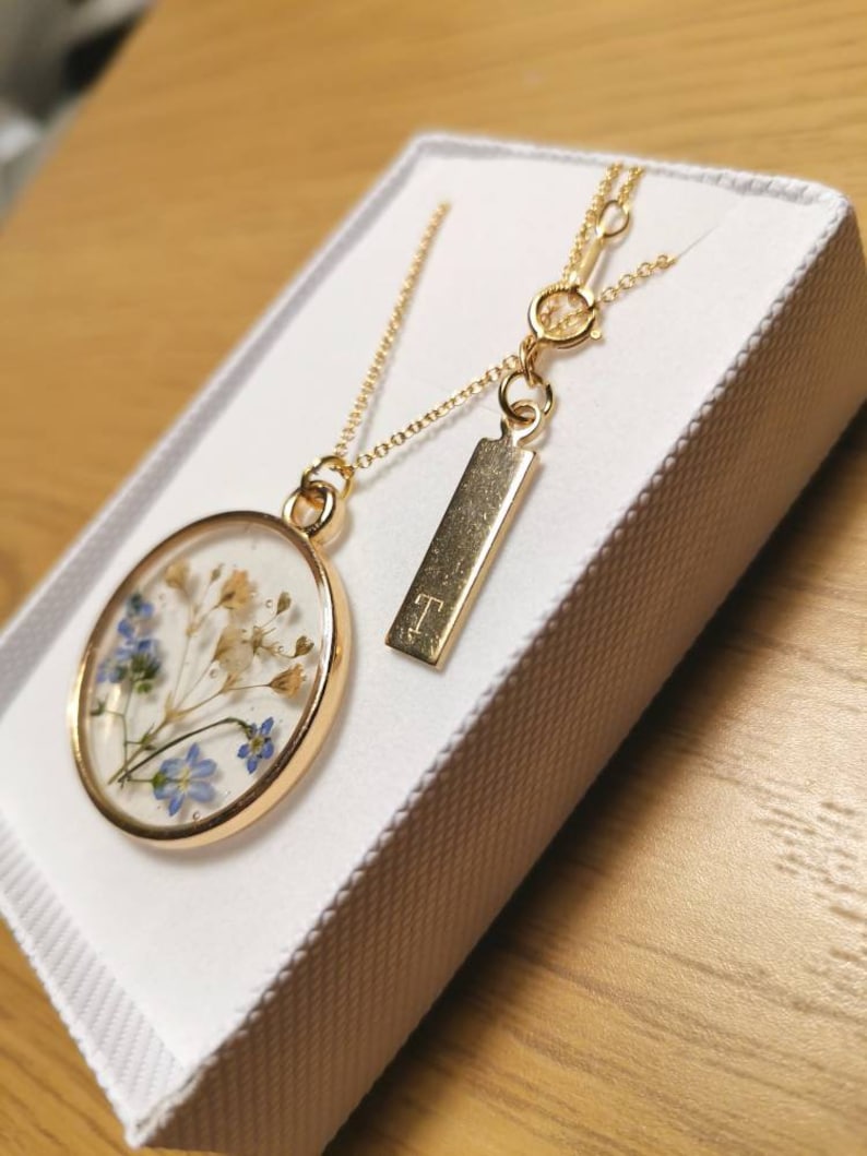 K&ouml;nnte beinhalten: Eine goldene Halskette mit einem runden Anh&auml;nger, der gepresste blaue und wei&szlig;e Blumen enth&auml;lt. Der Anh&auml;nger ist an einer zarten goldenen Kette befestigt. Die Halskette enth&auml;lt au&szlig;erdem einen kleinen goldenen Anh&auml;nger mit dem eingravierten Buchstaben "T".