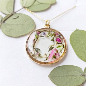 May include: A gold necklace with a round pendant featuring a delicate arrangement of pressed pink and purple flowers encased in clear resin.