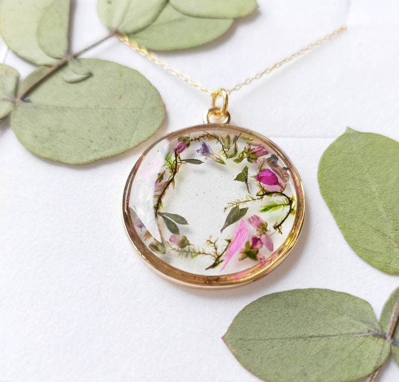 May include: A gold necklace with a round pendant featuring a delicate arrangement of pressed pink and purple flowers encased in clear resin.