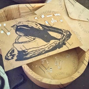 May include: A kraft paper card and envelope set featuring a black illustration of a skull. The card is inside a wooden bowl. The text "JML'26" is visible on the card. The words "CUSTOM DESIGNS" and "JODI LARSON" are printed on the card and bowl.
