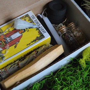 May include: A box containing a Rider-Waite tarot deck, a pendulum, incense, small glass bottles, sage, and charcoal discs. The tarot deck has a yellow cover with the words "The Rider Tarot Deck" and an image of "The Magician".